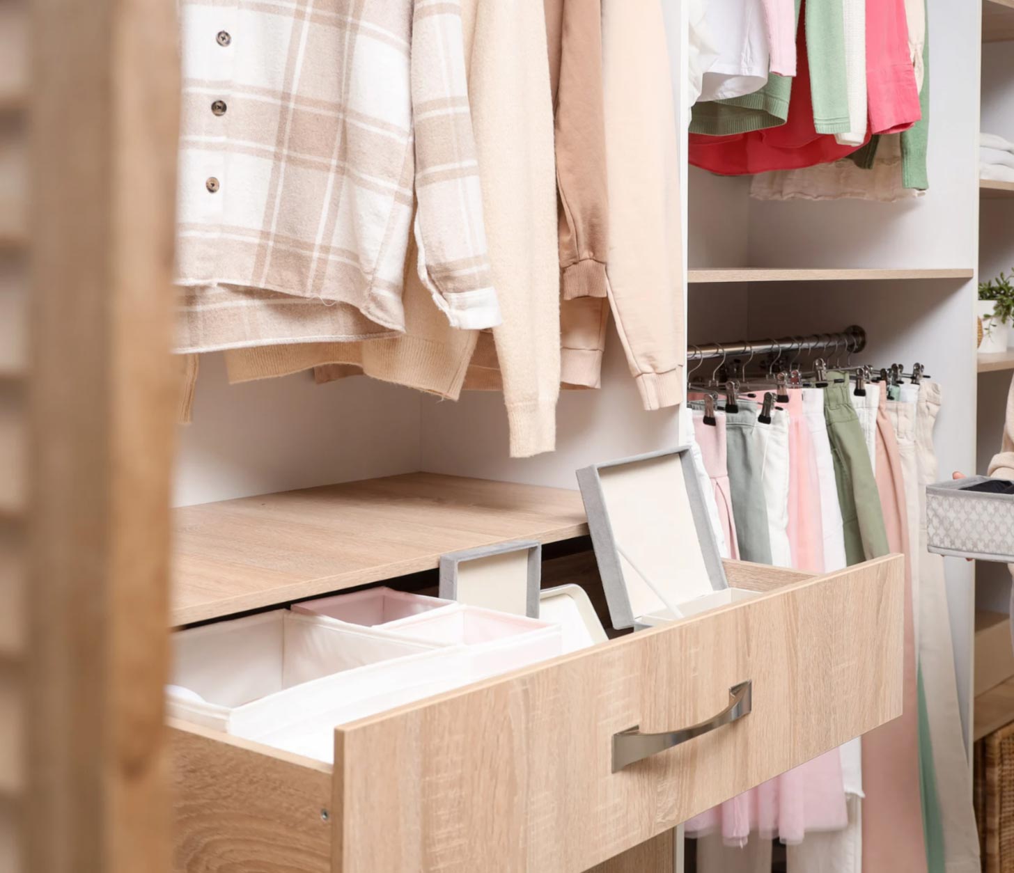 order-7 Open wooden closet drawer with organizers, below neatly hung pastel and neutral clothes on hangers.