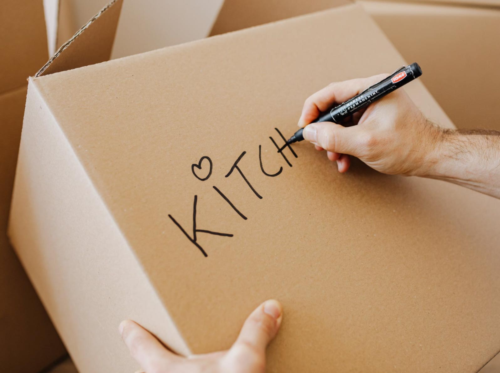 Hand writing "KITCHEN" on a cardboard moving box with a black marker; other boxes in the background.