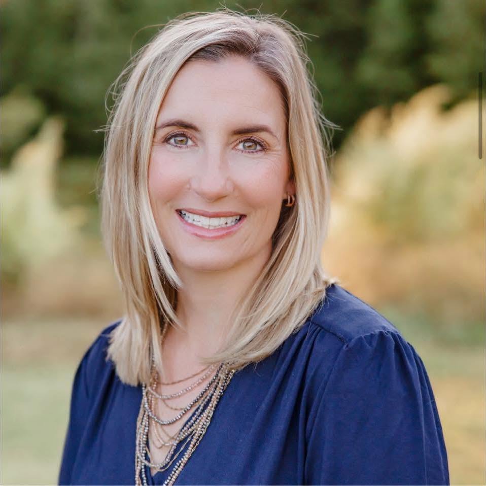 Smiling woman with blonde hair wearing a navy blouse, outdoors with greenery in the background.