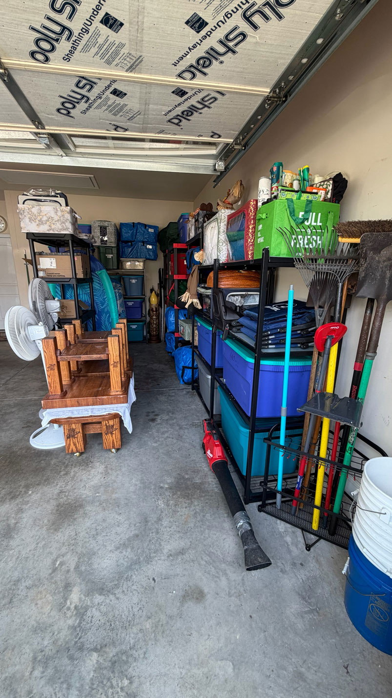 Organized garage with shelves holding bins, tools, and bags; wooden furniture and tools are on the left.
