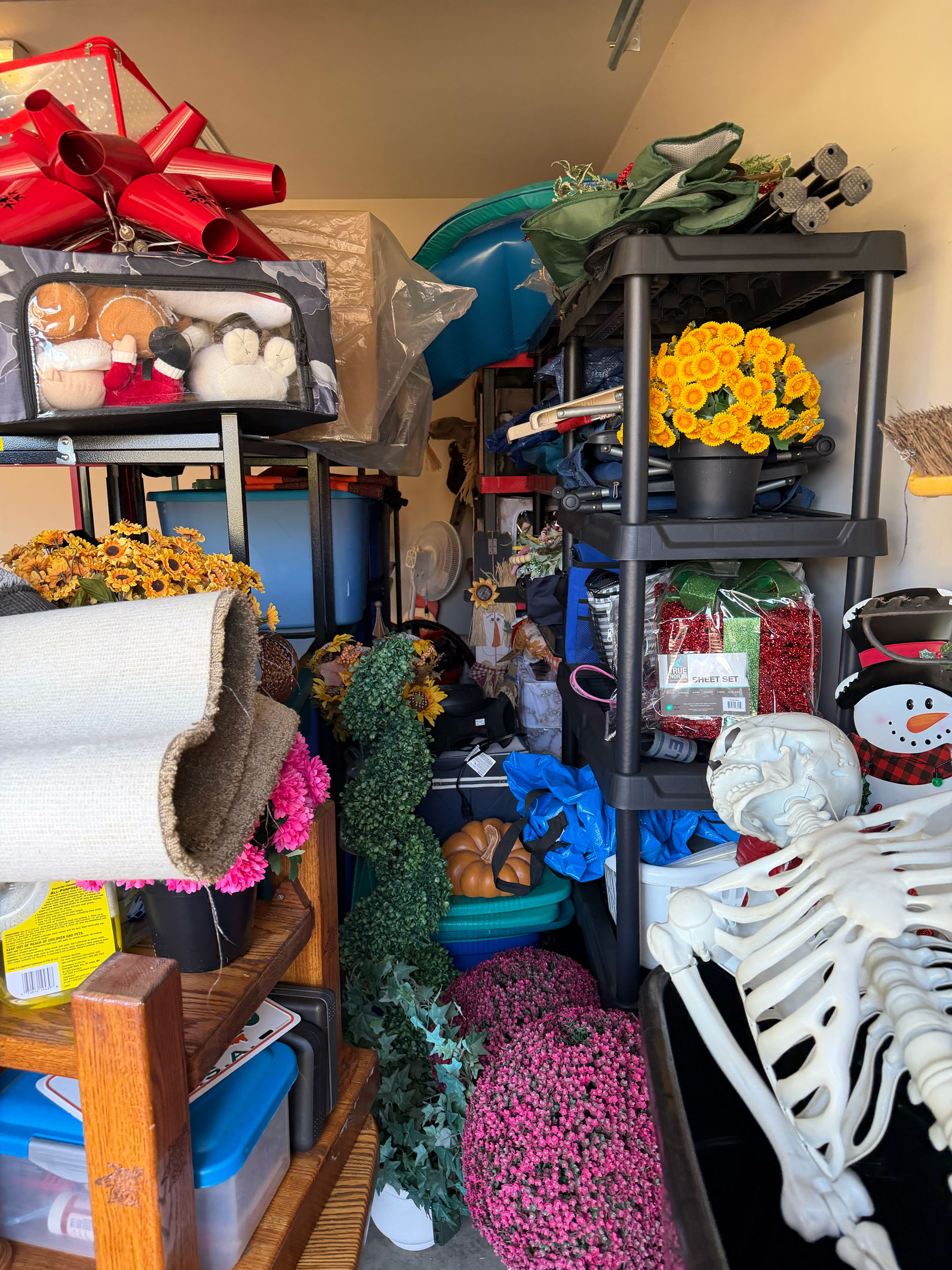Crowded storage room full of holiday and home decor, artificial flowers, plastic skeleton, and stacked shelving units.