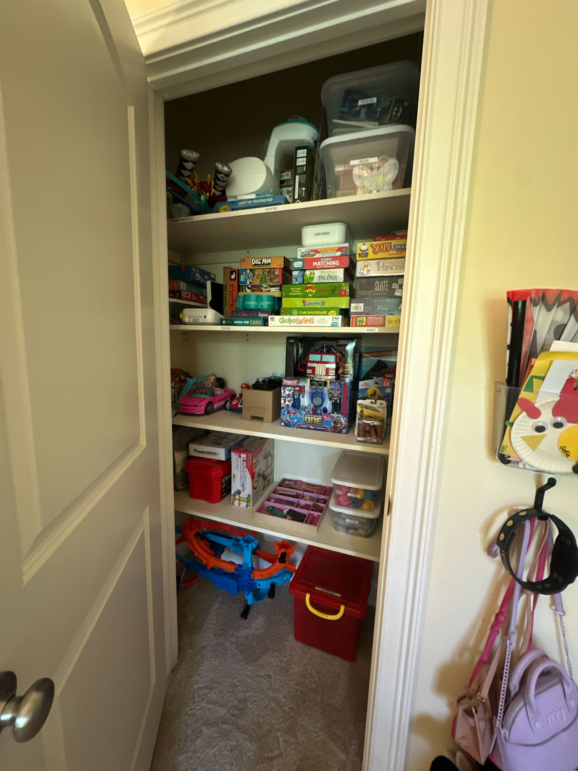 Closet shelves filled with board games, toys, and bins; door open, hooks with bags on the right.