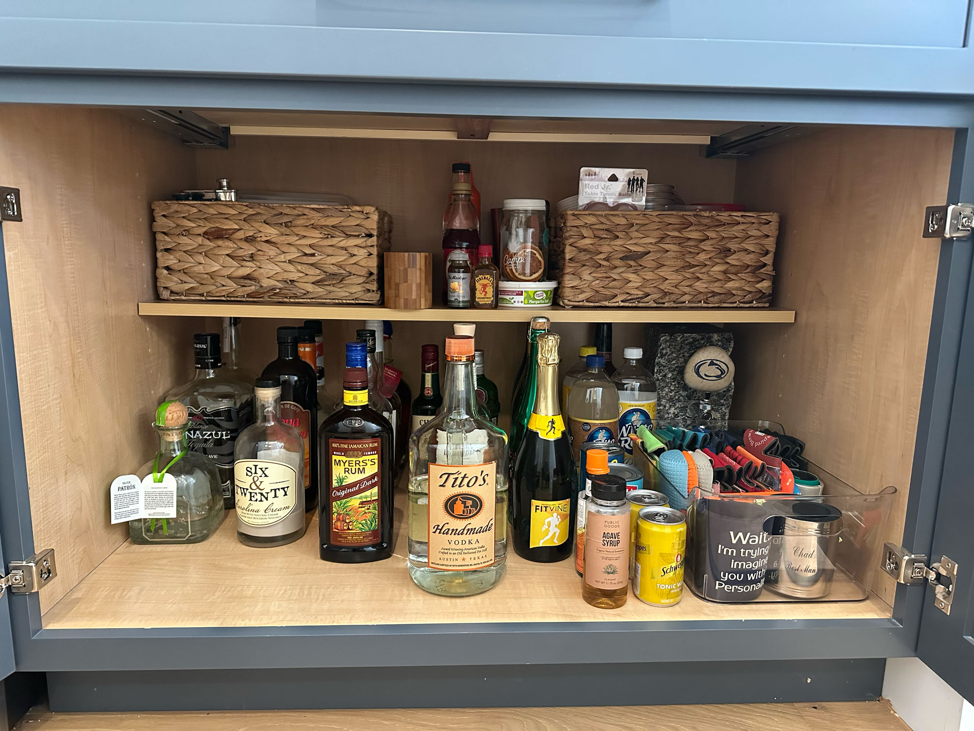 Cabinet filled with liquor bottles, mixers, mugs, glasses, and baskets on two wooden shelves.