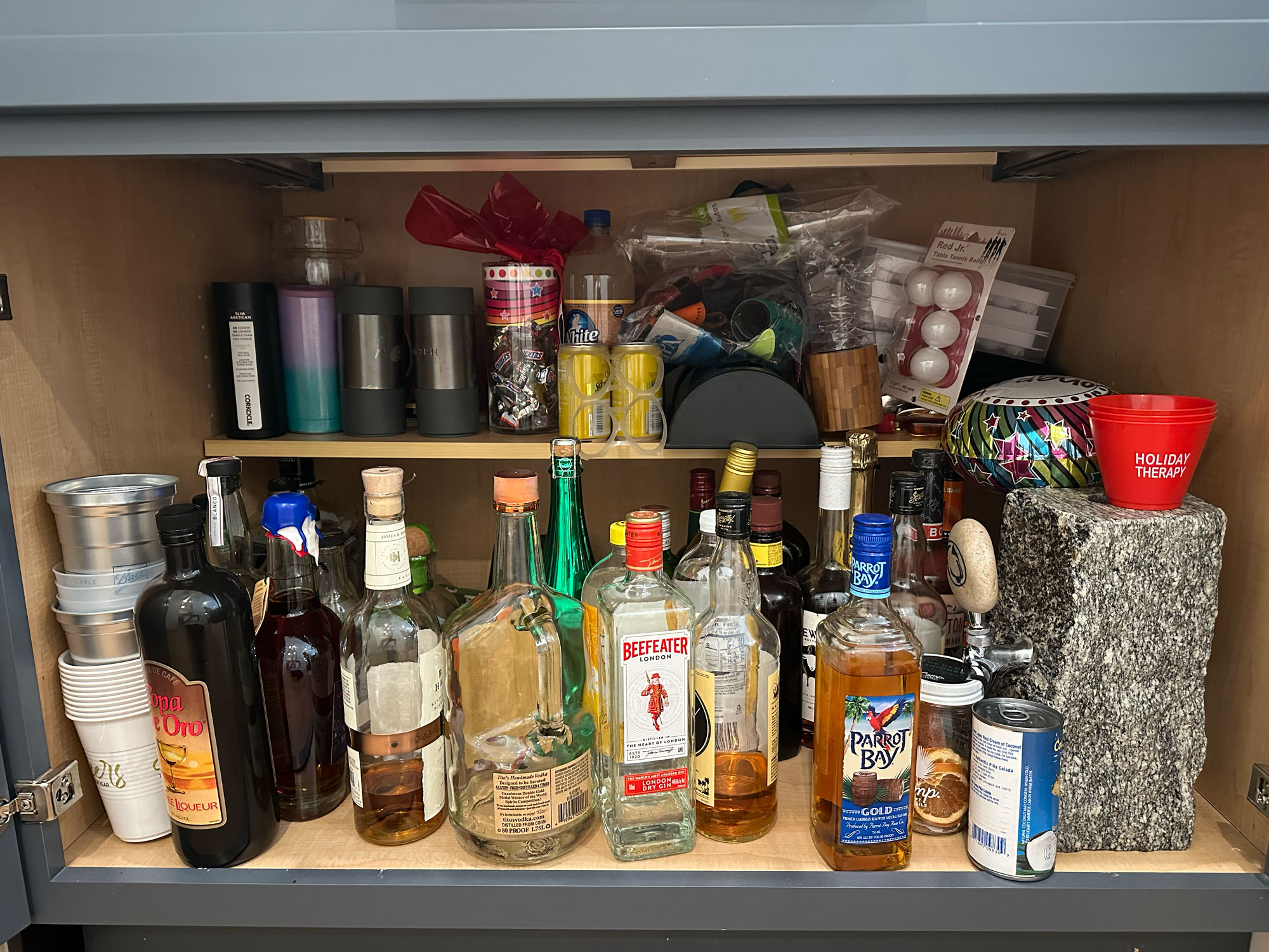 A cabinet with liquor bottles, cups, ping pong balls, party supplies, and cocktail accessories on two shelves.