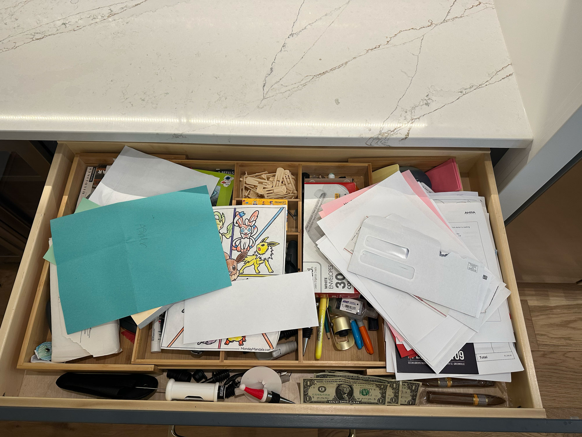 An open kitchen drawer contains papers, envelopes, pens, and a visible one-dollar bill.